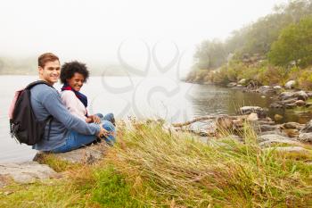 Hiking couple relaxing by the edge of a lake, looking to camera