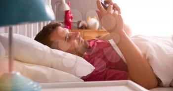 Man Lying In Bed Checking Messages On Mobile Phone