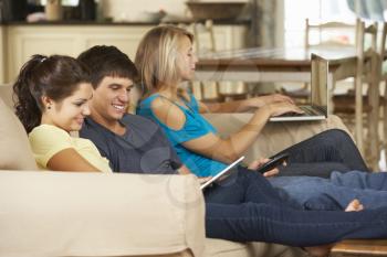 Three Teenagers  Sitting On Sofa At Home Using Mobile Phone, Tablet Computer And Laptop