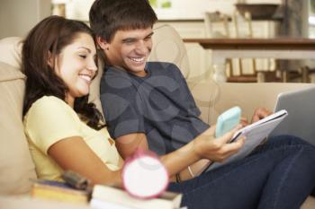 Teenage Boy And Girl Sitting On Sofa At Home Doing Homework Using Laptop Computer Whilst Holding Mobile Phone