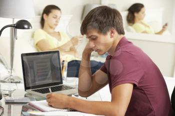 Teenage Boy Studying At Desk In Bedroom With Girl In Background