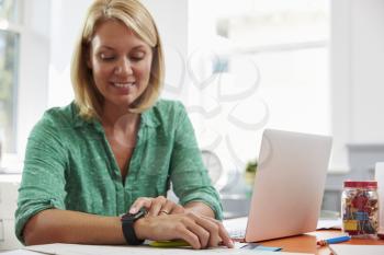 Woman Sitting At Desk In Home Office Looking At Smart Watch