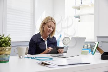 Female Doctor Sitting At Desk Using Digital Tablet In Office