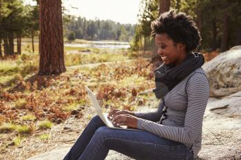 Black woman sitting on a rock in countryside using computer