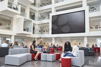 Students sit talking under AV screen in atrium at university