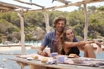 Young couple sitting at a table by the sea, look to camera