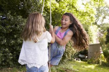 Two Girls Playing Together On Tire Swing In Garden