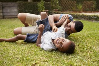 Black father and young son play lying on grass, low angle