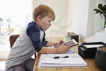 Boy Sits At Desk In Bedroom With Digital Tablet Doing Homework