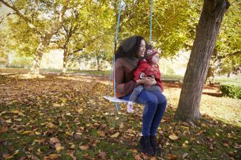 Mother With Daughter Playing On Tree Swing In Autumn Garden