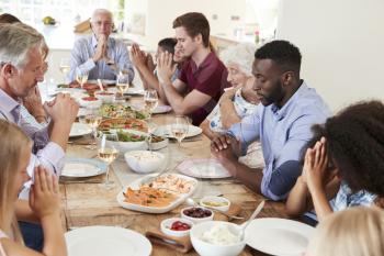 Multi-Generation Family And Friends Around Table Praying Before Meal At Party