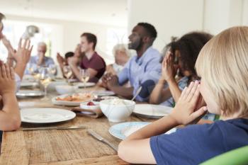 Multi-Generation Family And Friends Around Table Praying Before Meal At Party