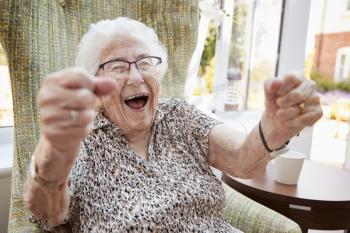 Portrait Of Excited Senior Woman Sitting In Chair In Lounge Of Retirement Home