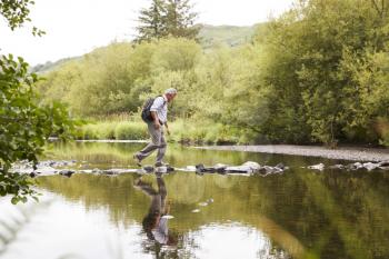 Senior Man Crossing River Whilst Hiking In UK Lake District