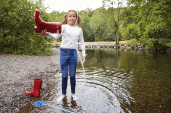 Teenage girl standing in a river wearing socks pours water out of her red wellington boot