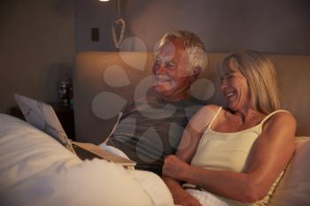 Senior Couple Wearing Pajamas Lying In Bed Looking At Laptop