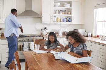 Daughters Sitting At Table Doing Homework As Father Cooks Meal