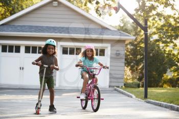 Sister With Brother Riding Scooter And Bike On Driveway At Home