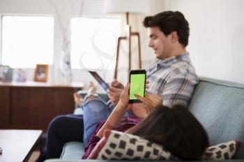 Couple Relaxing On Sofa At Home Using Mobile Phone And Digital Tablet