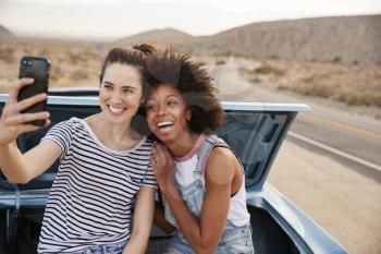 Two Female Friends Posing For Selfie Sitting In Trunk Of Classic Car On Road Trip