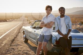 Portrait Of Two Male Friends Enjoying Road Trip Standing Next To Classic Car On Desert Highway