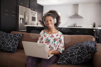 Portrait Of Woman Wearing Wireless Headphones Sitting On Sofa At Home Using Laptop