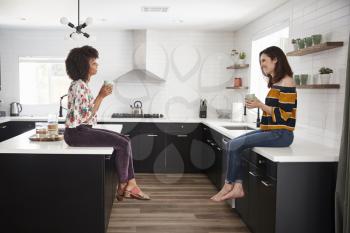 Two Female Friends Meeting For Coffee At Home Sitting On Kitchen Island