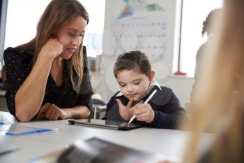 Young female teacher working with a Down syndrome schoolboy sitting at desk in a primary school classroom, selective focus