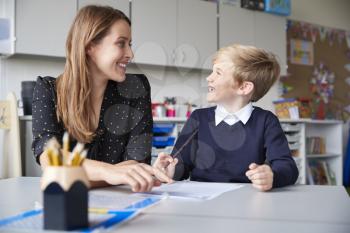 Young female primary school teacher and schoolboy sitting at a table working one on one, looking at each other smiling, front view, close up