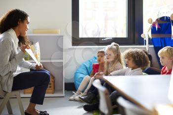 Side view of a female kindergarten teacher sitting on a chair showing a book to children in a classroom