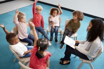 Young female school teacher reading a book to infant school children, sitting on chairs in a circle in the classroom raising hands to answer a question, elevated view