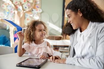 Female infant school teacher working one on one in a classroom using a tablet computer with a young mixed race schoolgirl, smiling at each other, close up