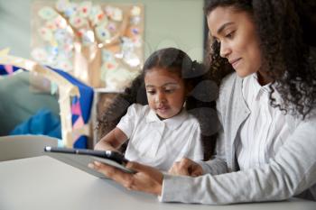 Close up of a young black schoolgirl sitting at a table in an infant school classroom learning one on one with a female teacher using a tablet computer, close up