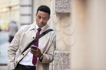 Young black businessman leaning on a wall in the street in London using smartphone, selective focus