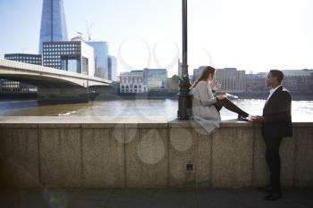 Two millennial colleagues take a break sitting on the embankment eating near London Bridge by the River Thames, backlit