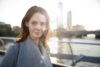 Young white woman wearing grey coat standing on Millennium Bridge, London, looking to camera smiling