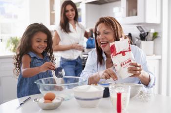 Young girl making a cake in the kitchen with her grandmother, mum stands watching, selective focus