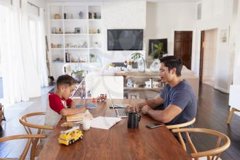 Hispanic father and son working opposite each other at the dining room table, concentrating