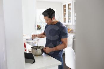 Millennial Hispanic man preparing cake mixture in kitchen, following a recipe on a tablet computer