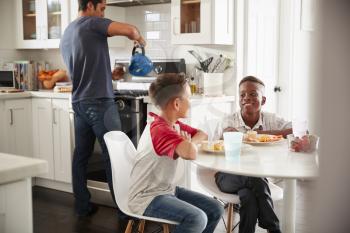 Two pre-teen male friends sit talking in kitchen at one boys house, dad in the background