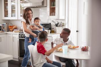 Mother holding baby stands in the kitchen talking with her son and his friend, over for a playdate