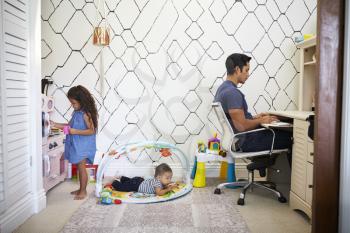 Dad sits working at a desk at home while his baby son and young daughter play in the room behind him