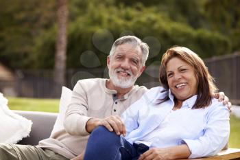 Senior couple sitting together on a seat in the garden smiling to camera, front view