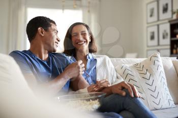 Young adult mixed race couple sitting on the sofa in their living room laughing and eating popcorn, selective focus