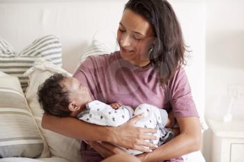 Young mixed race adult mother sitting on her bed and holding her baby son in her arms, waist up, close up