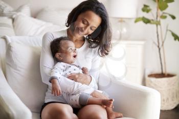 Young adult mother sitting in an armchair in her bedroom, holding her three month old baby son in her arms and looking down at him smiling, close up