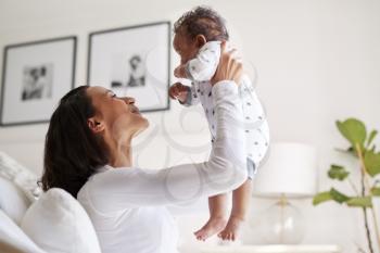 Happy mixed race young adult mother raising her three month old baby son in the air, smiling at him, close up, side view