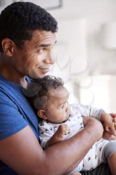 Close up of young adult black father sitting in an armchair holding his three month old baby son, side view, waist up, vertical