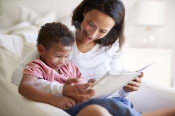 Close up of mixed race young adult mother sitting in an armchair reading a book with her three year old son on her knee, laughing, close up
