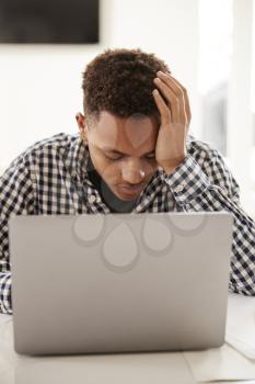 Depressed black teenage boy with head in hands using a laptop computer at home, vertical